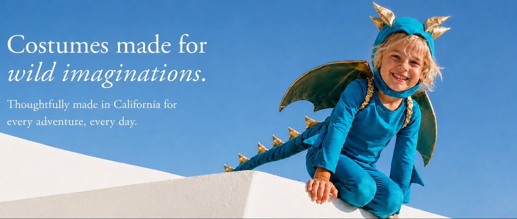A child wearing a blue dragon costume with green wings and gold horn details, sitting on a white wall against a clear blue sky. Text on the left reads, 'Costumes made for wild imaginations.