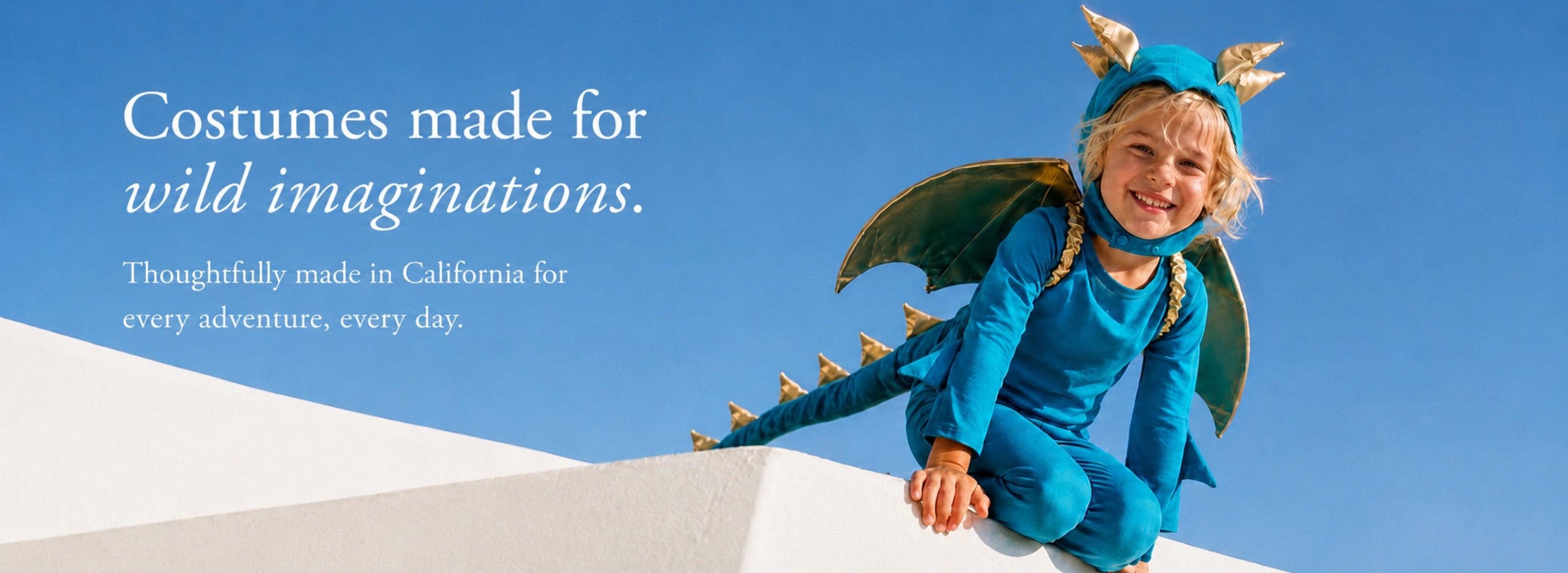 A child wearing a blue dragon costume with green wings and gold horn details, sitting on a white wall against a clear blue sky. Text on the left reads, 'Costumes made for wild imaginations.