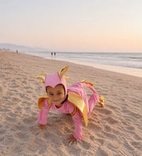 Toddler playing in a pink dragon costume with gold wings and horns on the sand